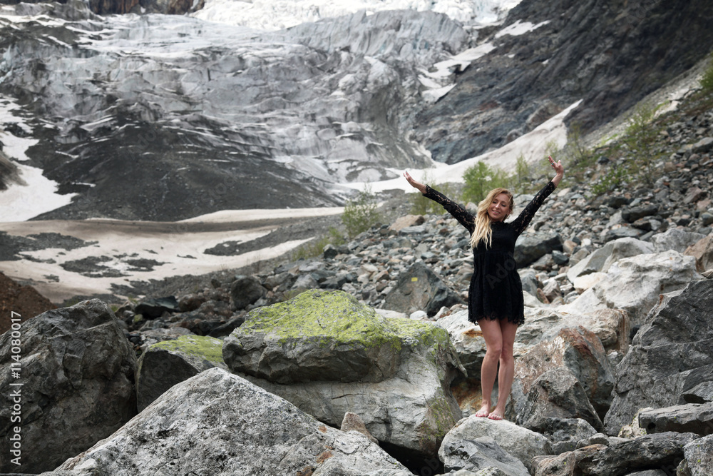 happy young blond woman in black dress among the huge stones on the glacier. Mestia