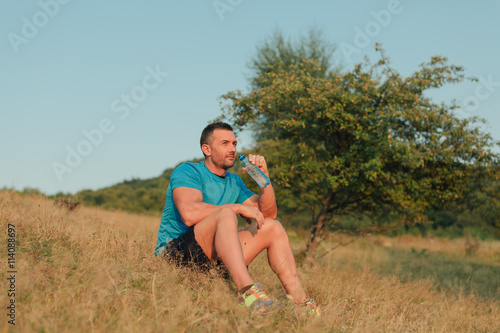 Athletic man in his 30s resting and drinking water from a bottle while sitting down outdoor