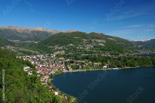 Swiss city of Caslano at the west side of the Lago di Lugano. Bare and snow covered mountains in the background. Densely forested hills spotted with houses steeply descend to the lake shore.