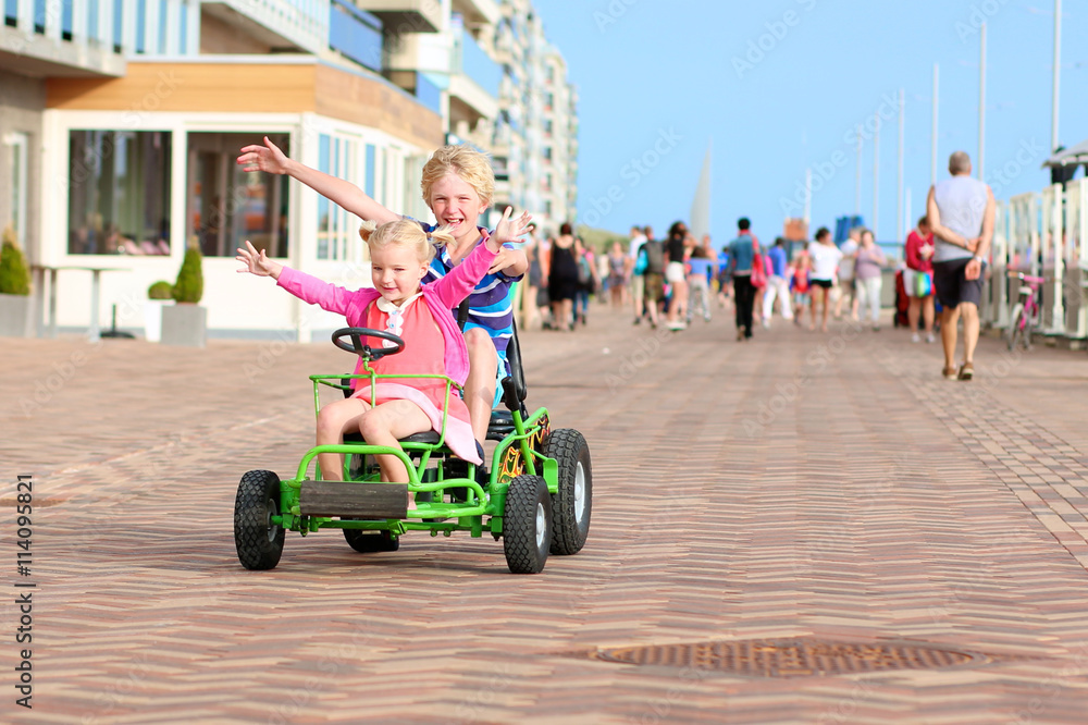Happy kids enjoying active holidays on the beach. Sportive boy riding pedal car together with his cute toddler sister along the promenade on a summer day at sunset. Happy kids enjoying active holidays on the beach. Sportive boy riding pedal car together with his cute toddler sister along the promenade on a summer day at sunset.