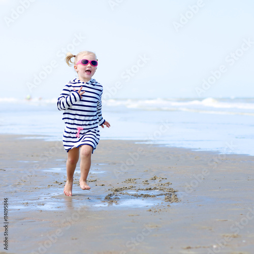 Cute active child wearing pink sunglasses playing and running on wide sandy beach. Happy little girl enjoying summer holidays on a sunny day. Family with young kids on vacation at the North Sea coast.