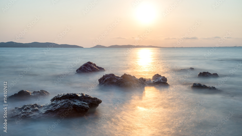 Romantic atmosphere in peaceful sunset at sea. Big boulders sticking out from smooth wavy sea.