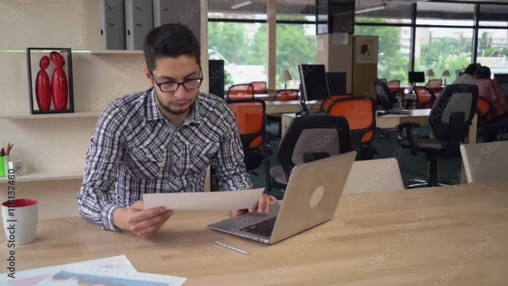 Businessman wearing in casual shirt. Male looking on the computer screen. Loft in contemporary company with many wooden table device and technical equipment. He could be marketing manager or Businessman wearing in casual shirt. Male looking on the computer screen. Loft in contemporary company with many wooden table device and technical equipment. He could be marketing manager or