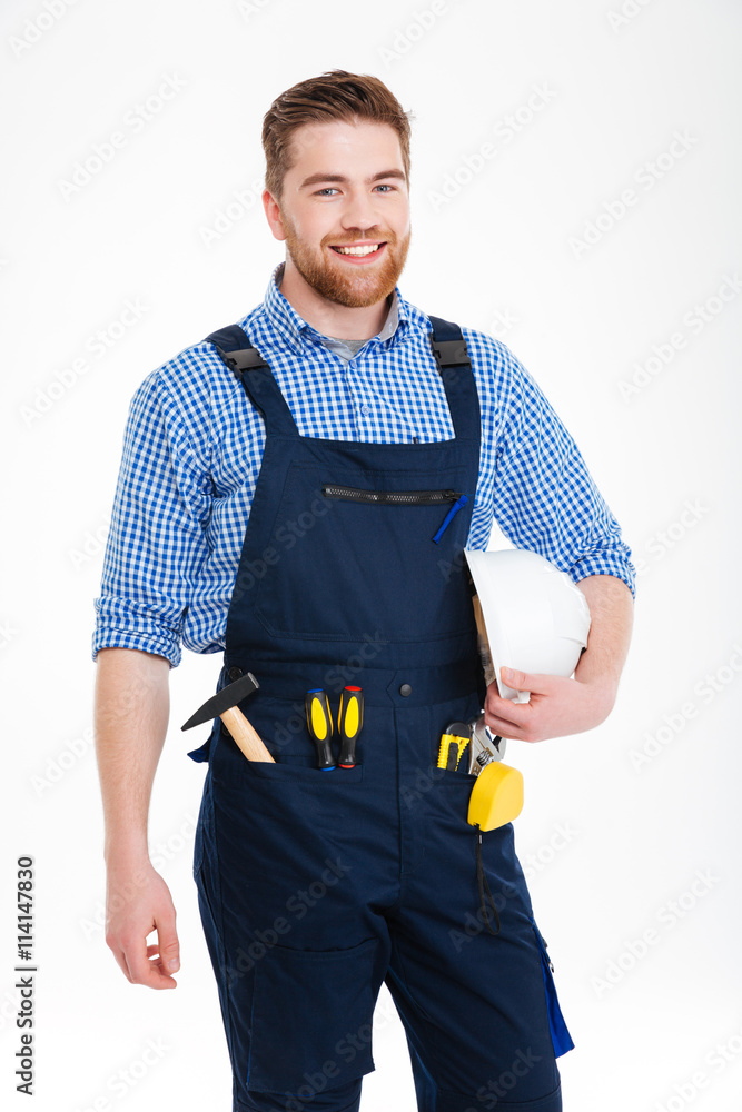 Cheerful handsome young builder in overall standing and holding helmet Cheerful handsome young builder in overall standing and holding helmet
