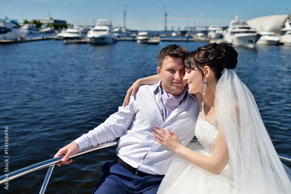 Wedding couple is hugging on a yacht. Beauty bride with groom. Beautiful model girl in white dress. Man in suit. Female and male portrait. Woman with lace veil. Cute lady and guy outdoors Wedding couple is hugging on a yacht. Beauty bride with groom. Beautiful model girl in white dress. Man in suit. Female and male portrait. Woman with lace veil. Cute lady and guy outdoors