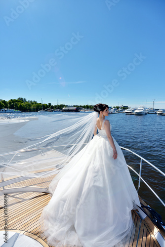 Beauty bride in bridal gown with lace veil on a yacht. Beautiful model girl in a white wedding dress. Female portrait on the nature. Woman with hairstyle. Cute lady outdoors
