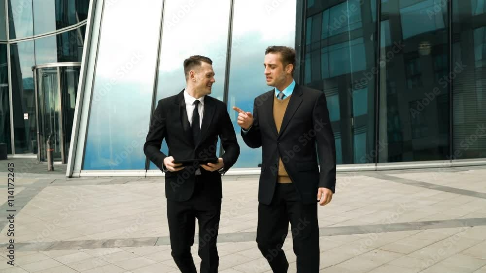 Two young successful businessmen easy communicating and discussing about something fun outdoor. Midday break. Tablet PC in hand. Glass business centre building at the background. Teal and Orange style Two young successful businessmen easy communicating and discussing about something fun outdoor. Midday break. Tablet PC in hand. Glass business centre building at the background. Teal and Orange style