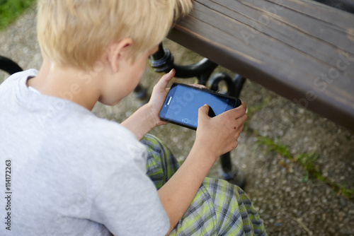 boy intently playing games on smartphone sittng on bench outside, summertime photo