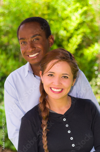 Beautiful young interracial couple in garden environment, embracing and smiling happily to camera