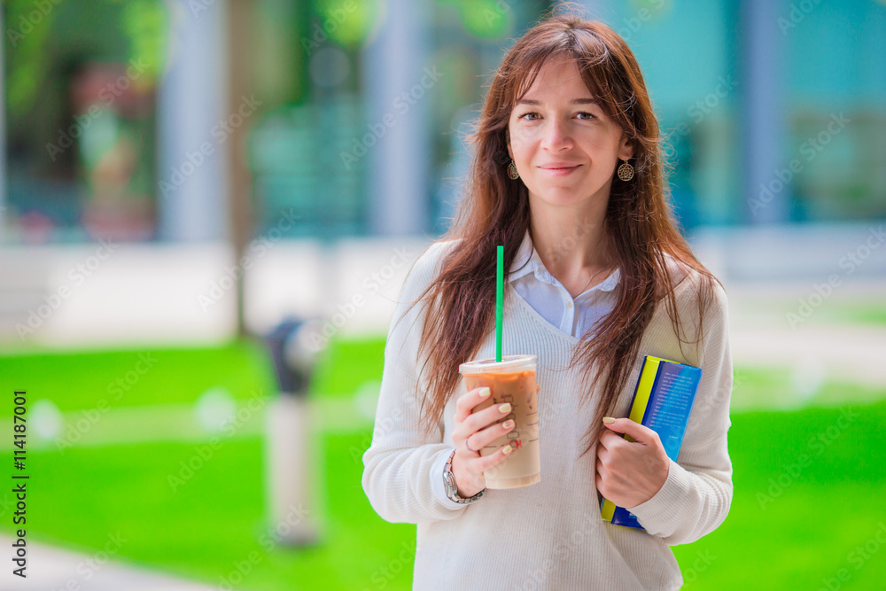 Happy young student girl with a coffee-to-go, walking in a summer park and holding books for reading.