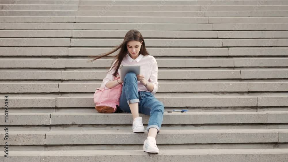 Young beautiful stylish girl sits on steps and holding hands in the tablet. Girl student communicates in social networks with friends.