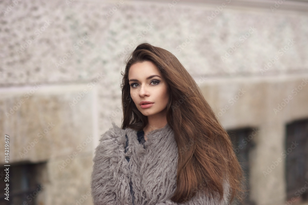 Fashion style portrait of young beautiful elegant woman in black dress and gray fur coat walking at city street