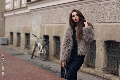 Fashion style portrait of young beautiful elegant woman in black dress and gray fur coat walking at city street