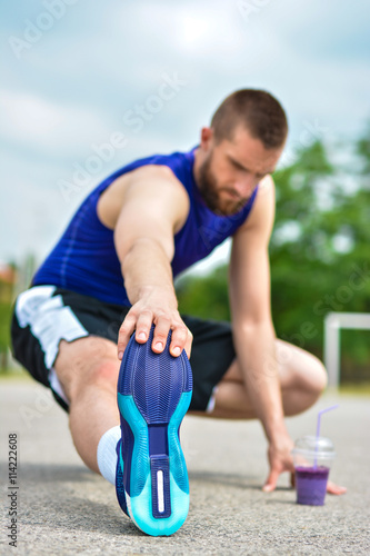 Athletic sportsman with a bottle of detox smoothie sitting and doing warming stretching exercise in the park