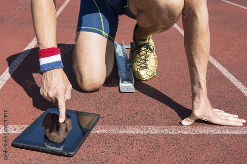American athlete in gold running shoes crouching at the starting line of a running track wearing USA colors wristbands using his tablet