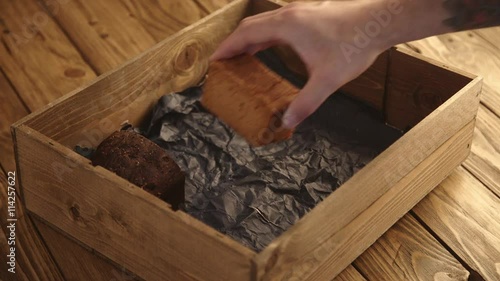 Baker with tattooed hand carefully puts different samples of mixed healthy breads from vegetable and fruits inside rustic wooden box with craft paper on old brushed table for presentation. photo