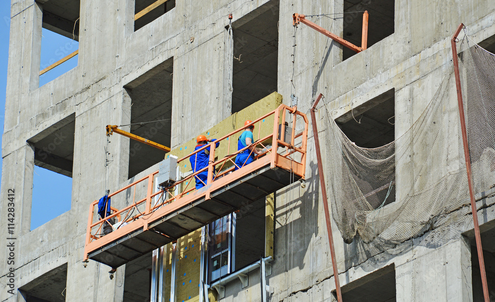 Отделка фасада строящегося высотного дома: рабочие в люльке монтируют панели утеплителя