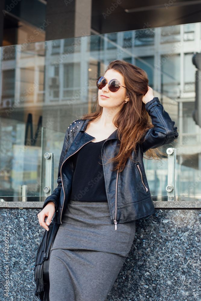Fototapeta premium Stylish young woman in leather jacket and sunglasses posing outdoor 