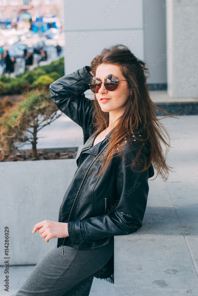 Stylish young woman in leather jacket and sunglasses posing outdoor 