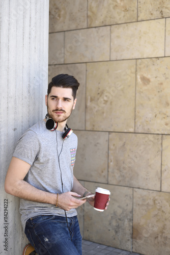 Young man holding a smart phone and coffe with headphones around