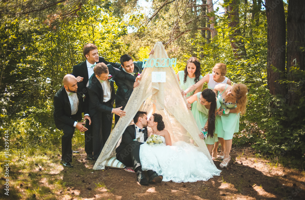Full length portrait of newlywed couple having fun with bridesmaids and groomsmen in green sunny park. Friends near hand made tent