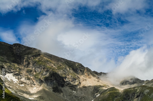 Rocky Mountaintop with bits of snow and grass in front of cloudy blue sky