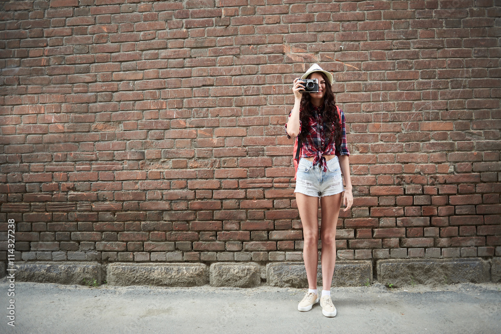 Portrait of a girl tourist in a hat taking pictures on the background wall red brick building