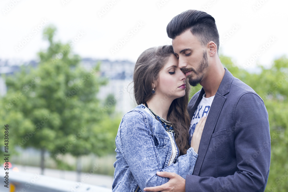 Young couple in love outdoors, in the city near a group of trees. She is touching his chest, very close to him. Young couple in love outdoors, in the city near a group of trees. She is touching his chest, very close to him.