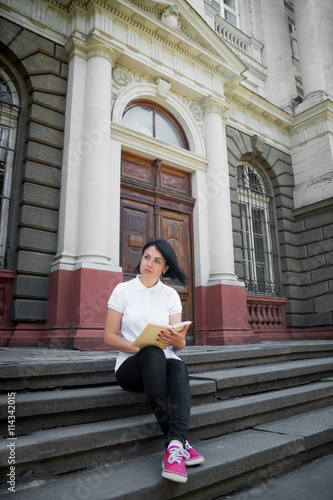 Woman is reading book. Girl with book in white polo, black pant and red shoes is sitting on stairs near university at campus and learning, studying. teenager sitting outdoors with open book