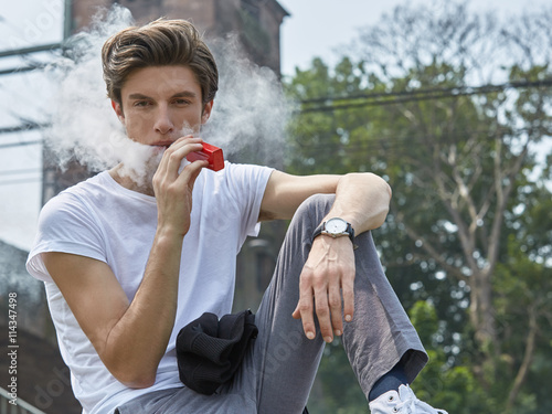 Junger Mann in T-Shirt und Jeans sitzt e-Zigarette rauchend an einer Bahnstrecke und bläst lässig eine weiße Dampfwolke.