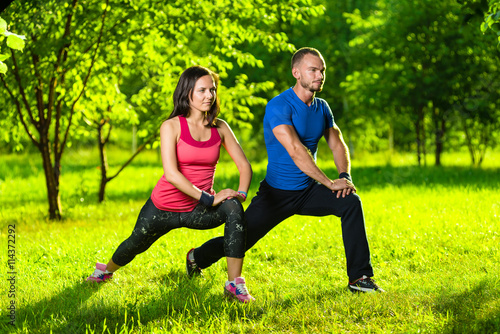 Man and woman doing stretching exercises at summer park. Young couple exercising and stretching muscles before sport activity - outdoor in nature