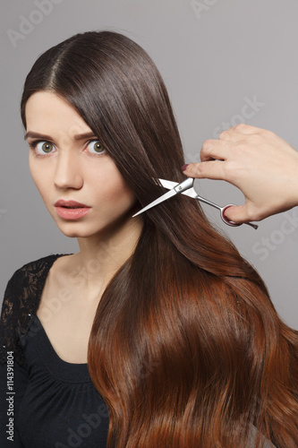 Hairdresser cutting gorgeous hair with hairdressing scissors over grey background. Portrait of frightened woman with modern hairstyle in studio.