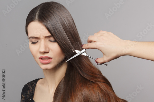 Hairdresser cutting gorgeous hair with hairdressing scissors over grey background. Young woman looking frightened in studio.