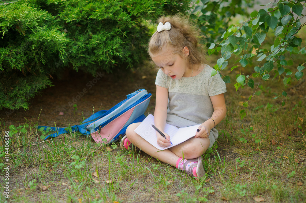 First grader sits having crossed legs under a tree and does homework. First grader sits having crossed legs under a tree and does homework.