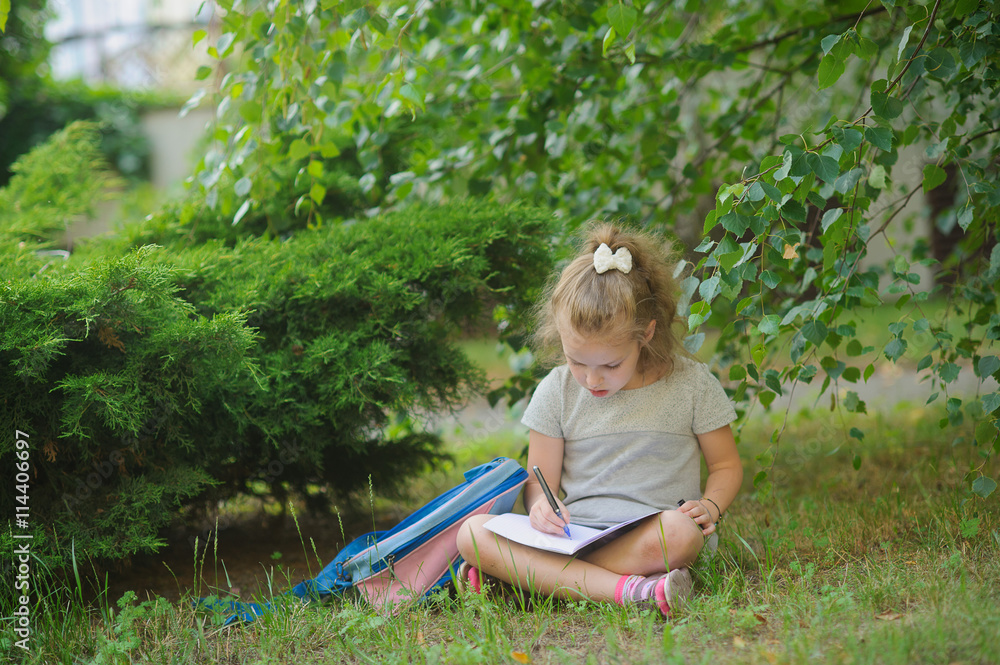 Little schoolgirl sits having crossed legs under a tree and does homework.