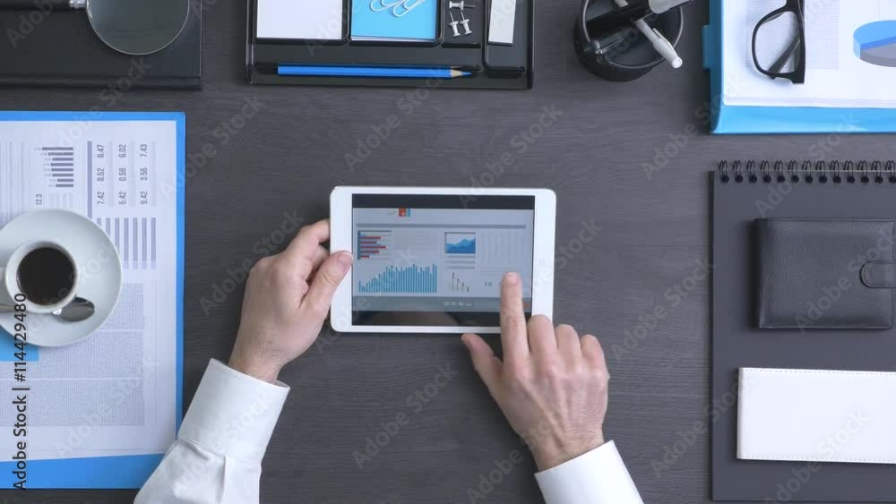 Businessman working at office desk and using a touch screen tablet, he is checking financial reports and using mobile apps Businessman working at office desk and using a touch screen tablet, he is checking financial reports and using mobile apps