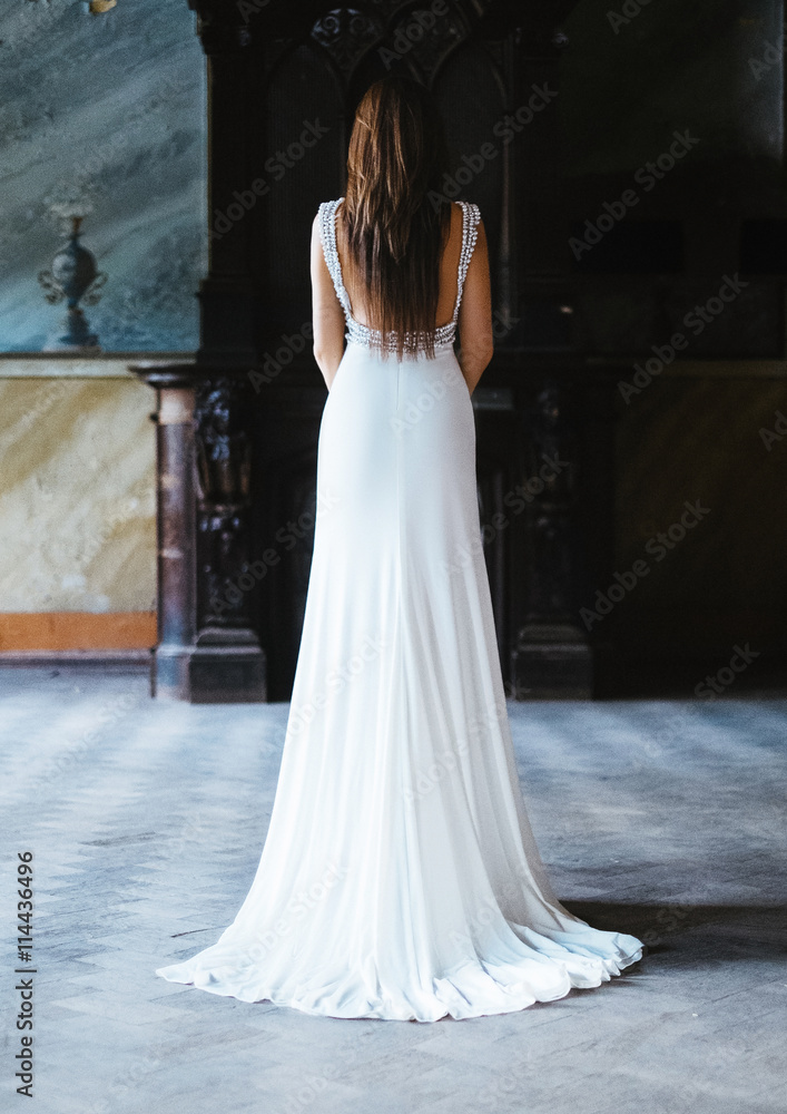 Fototapeta premium the effect of film photography. Beautiful sexy woman with long dark hair. Dressed in a white long dress with a train at the floor. Posing in an abandoned old castle on a background of fire.