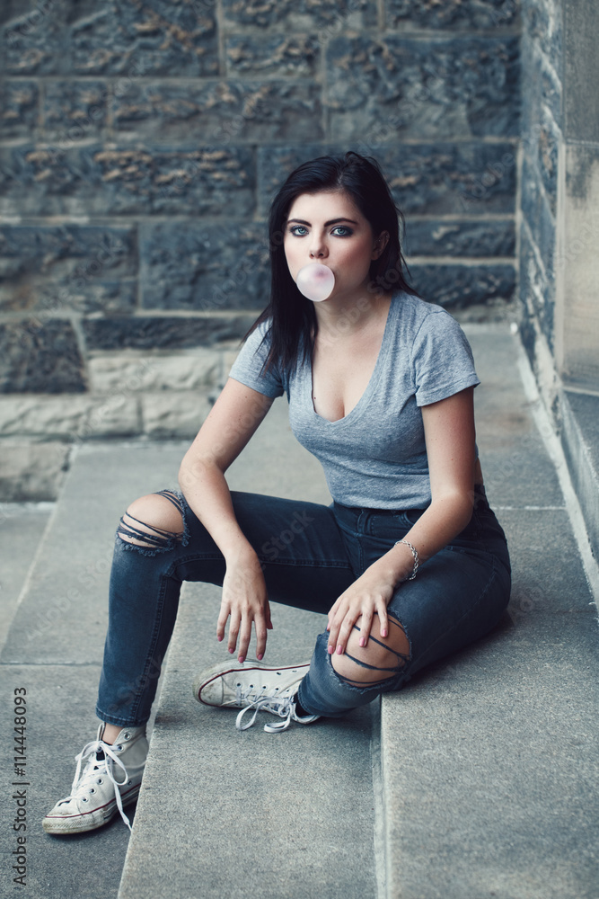 Portrait of white Caucasian beautiful young brunette girl, light blue green  eyes in ripped jeans, t-shirt, keds. Teenager blowing bubble gum looking in  camera. Toned with Instagram filters, lifestyle Stock Photo