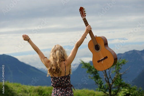Junge Frau streckt Arme mit Gitarre hoch vor Alpenpanorama