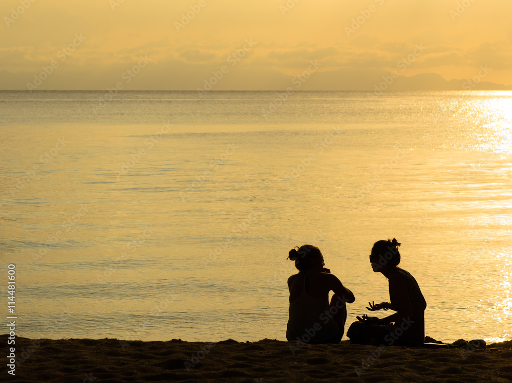Silhouette of two women together sitting and talking on beach.