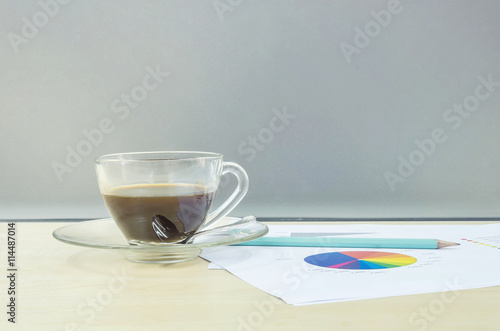 Closeup black coffee in transparent cup of coffee with work paper and pencil on blurred wooden desk and frosted glass wall textured background , work concept by coffee work paper and pencil