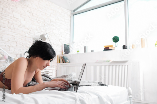 Woman typing on laptop in bedroom free space. Nude brunette lying on bed and working on laptop in her bedroom. Typing brunette in bra