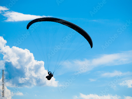 Dark paraglide silhouette on background of blue summer sky and white clouds. Adrenalin sport theme.