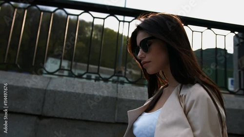 Confident young brunette woman in white jacket and black sunglasses walking down stairs in the city