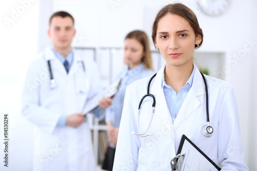 Young beautiful female doctor smiling  on the background with patient and his doctor in hospital. High level and quality medical service concept.