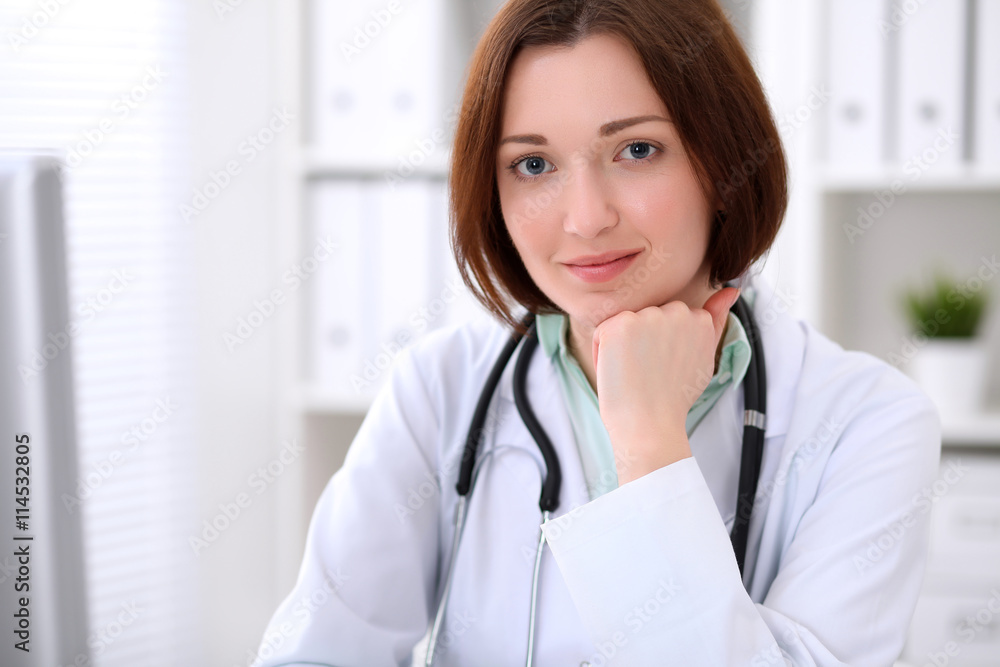 Young brunette female doctor sitting at a desk and working on the computer at the hospital office. Health care, insurance and help concept. Physician ready to examine patient Young brunette female doctor sitting at a desk and working on the computer at the hospital office. Health care, insurance and help concept. Physician ready to examine patient