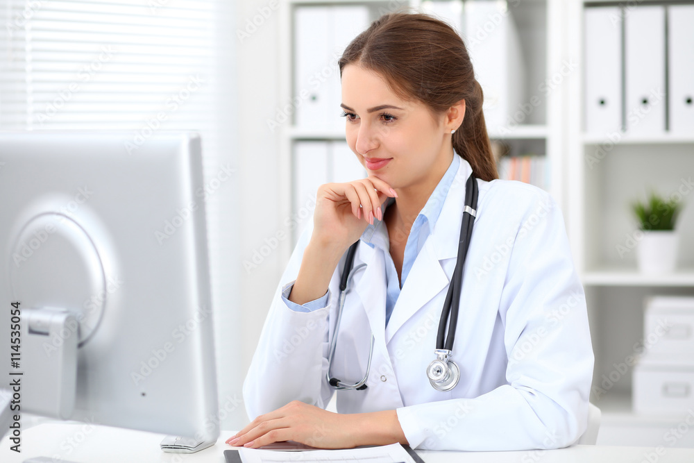Young brunette female doctor sitting at the table and working at hospital office. Health care, insurance and help concept. Physician ready to examine patient Young brunette female doctor sitting at the table and working at hospital office. Health care, insurance and help concept. Physician ready to examine patient