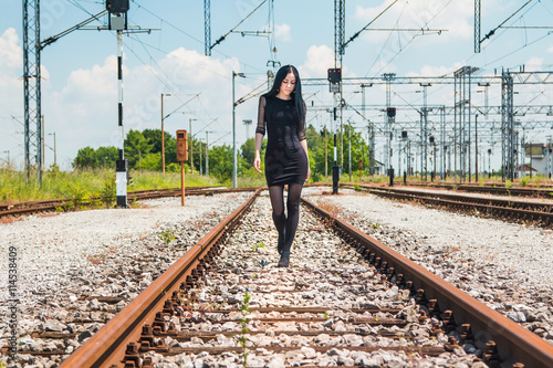  Young beautiful girl in black dress and nylons walking down rail tracks, cargo wagons in background 