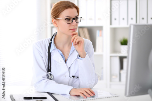 Young brunette female doctor sitting at the table and working by computer at hospital office. Doctor is in trouble with diagnosis. Health care, insurance and help concept.