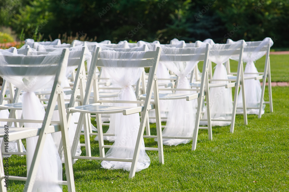 White decorated chairs on a green lawn. Chairs set in rows for the wedding ceremony. They are decorated for the festive event. Chairs are on the green lawn outside.